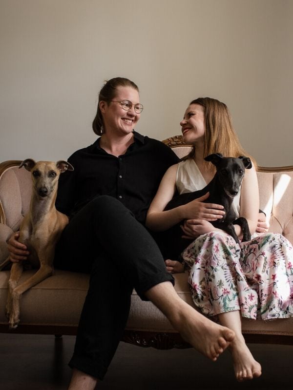 Owners of Heili Bridal, Valentin and Iida are sitting on a bluch rococoo couch. They smile and look at each others and both are holding their family whippets, Monty and Charlie, on lap.